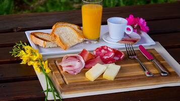 A picnic setup with bread, cheese, ham, and salami on a wooden board, a glass of orange juice, a cup, and cutlery on a white tray.