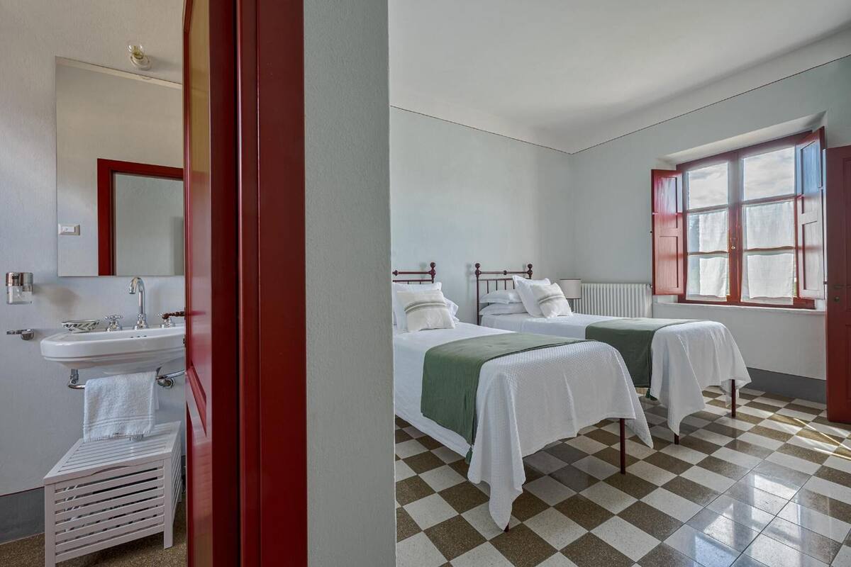A room with two beds, a window with red shutters, a white sink, and a mirror.