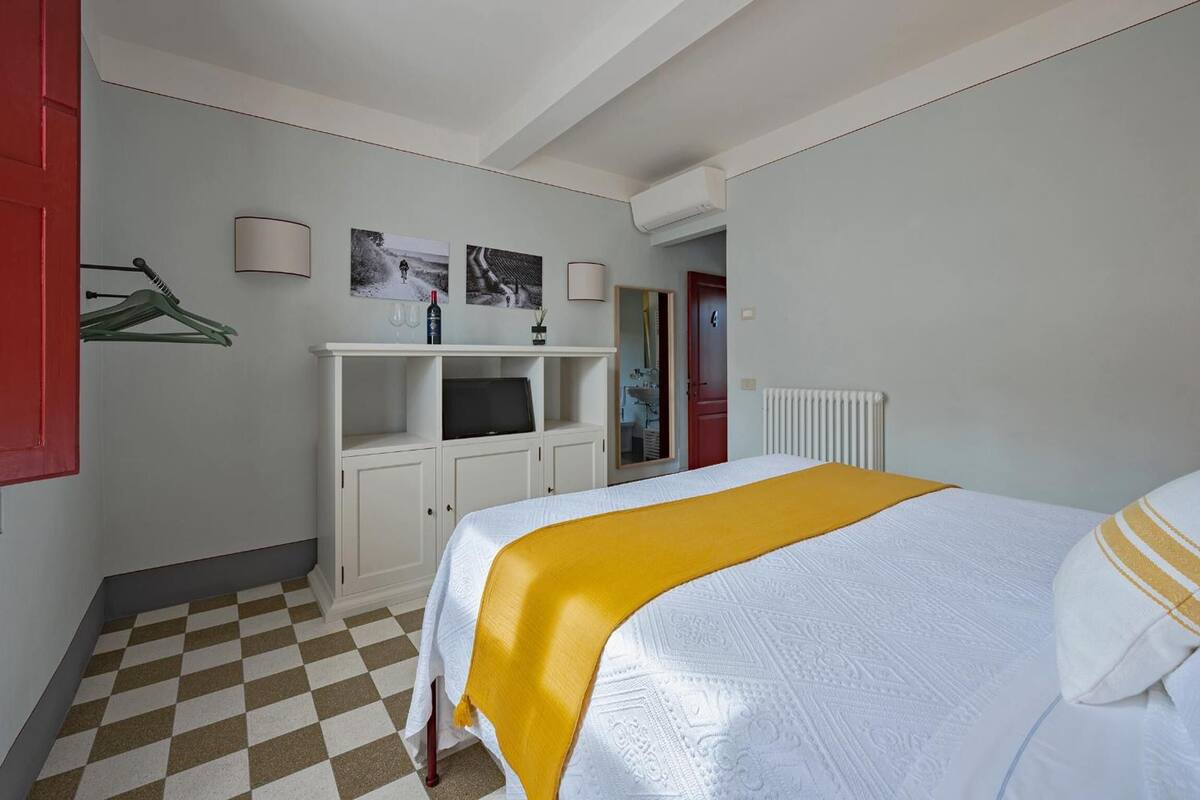 A hotel room with a checkered floor, a bed with a yellow blanket, a white wardrobe, a TV on a white cabinet, and a red door.