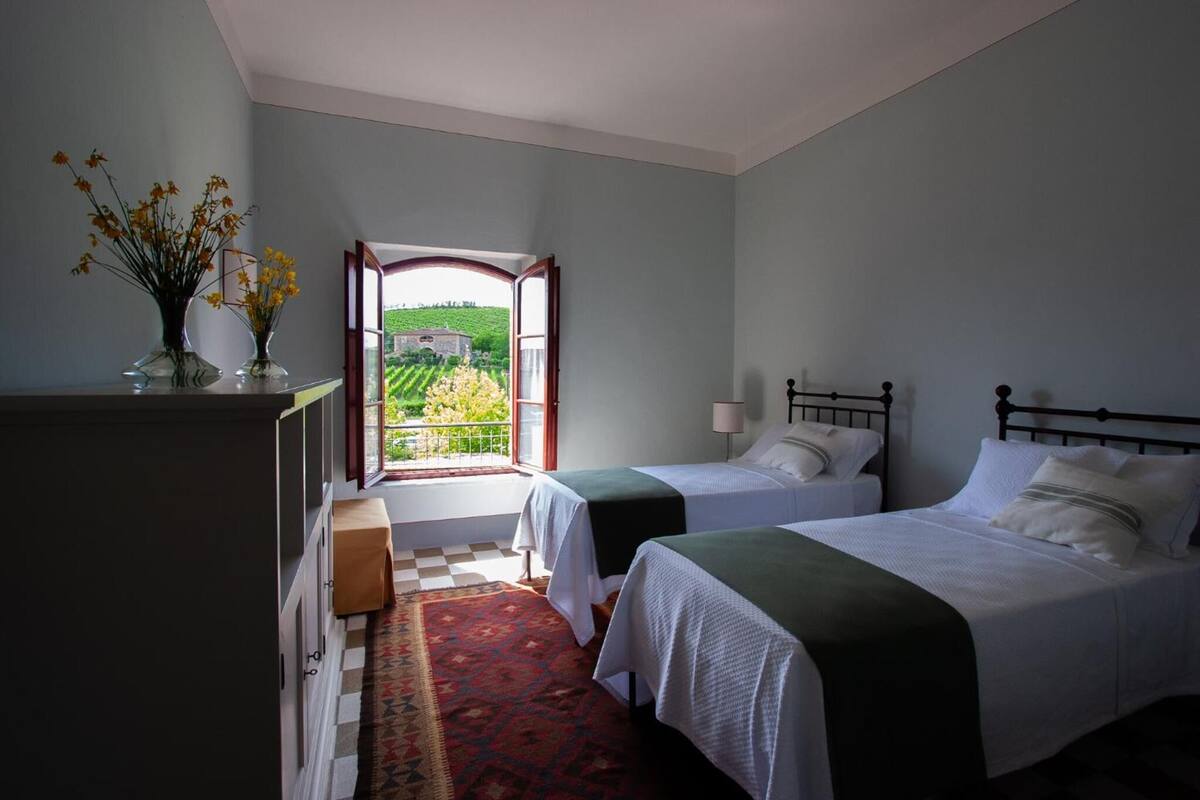 A bedroom with two beds, a window with a view of a green landscape, and a small table with a vase of flowers.