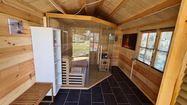 Interior de una cabaña de madera con un refrigerador blanco, un banco de madera y una puerta de vidrio que da a un espacio exterior.