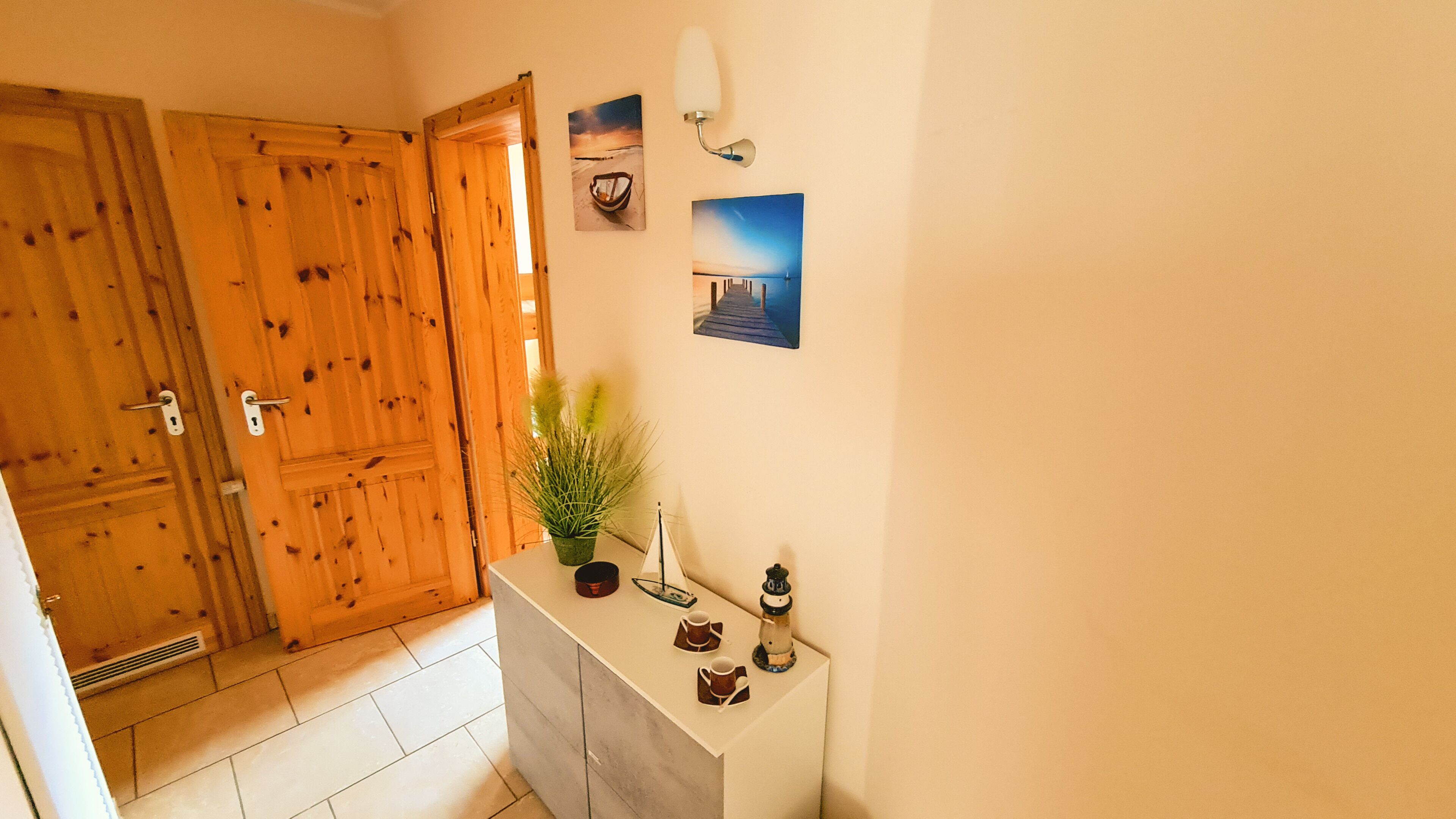 Une pièce avec des portes en bois, un sol carrelé, une petite console blanche ornée d’une plante, deux tableaux encadrés et un luminaire fixé au mur.
