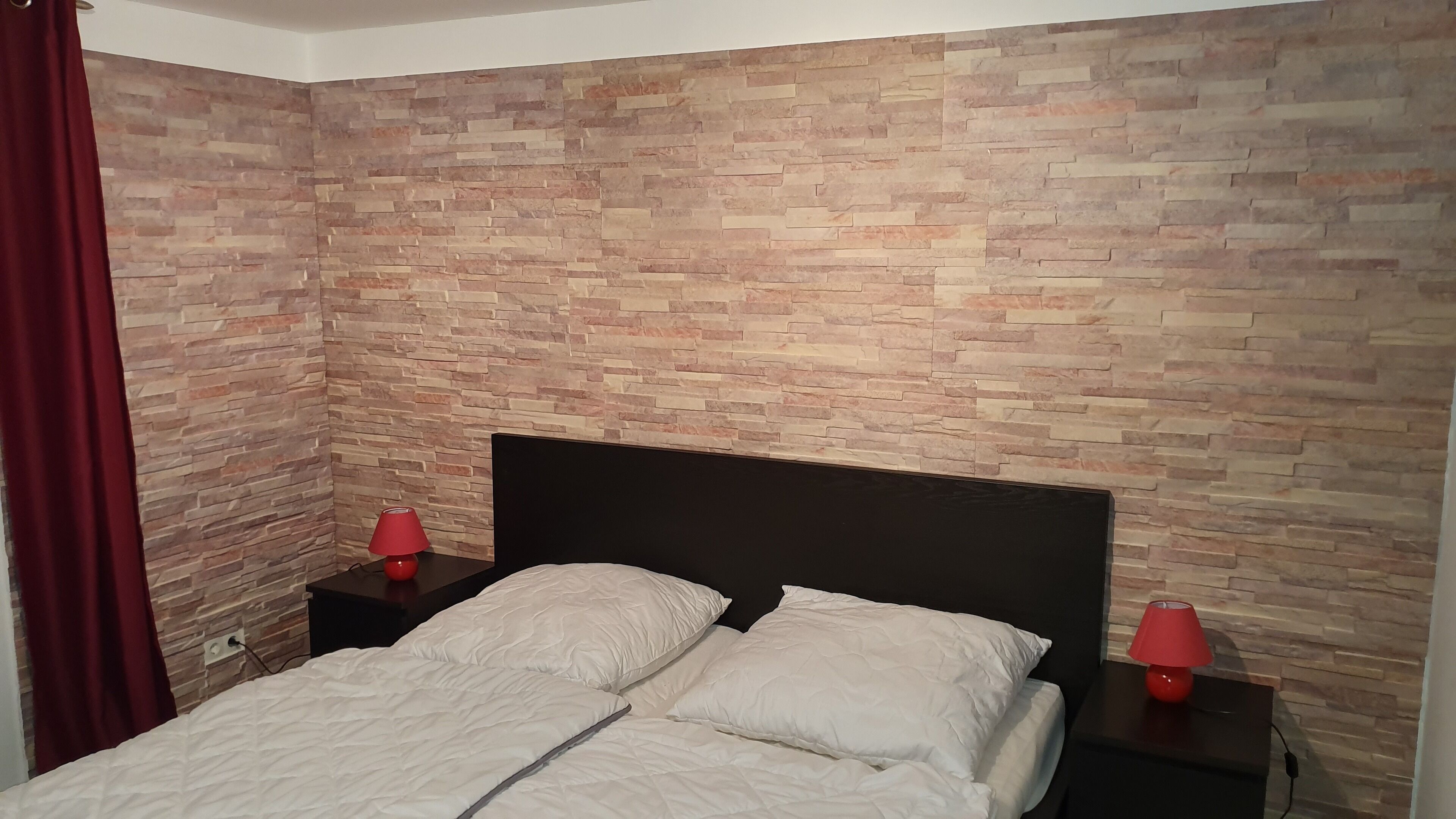 Un dormitorio con pared de ladrillo, cabecera negra, dos almohadas blancas y dos lámparas de noche rojas.