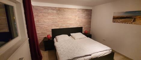 Un dormitorio con una cama, dos mesitas de noche, una ventana con cortinas, una pared con diseño de ladrillo y un cuadro en la pared.