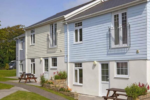 A row of two-story houses with white and light blue siding, each with a balcony and a small garden area with flowers. - Atlantic Lodge (Newquay)