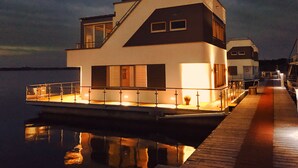 A modern houseboat with a flat roof, large windows, and a dock extending into calm water.