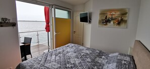 A bedroom with a bed, a TV, a painting, and a view of the sea.