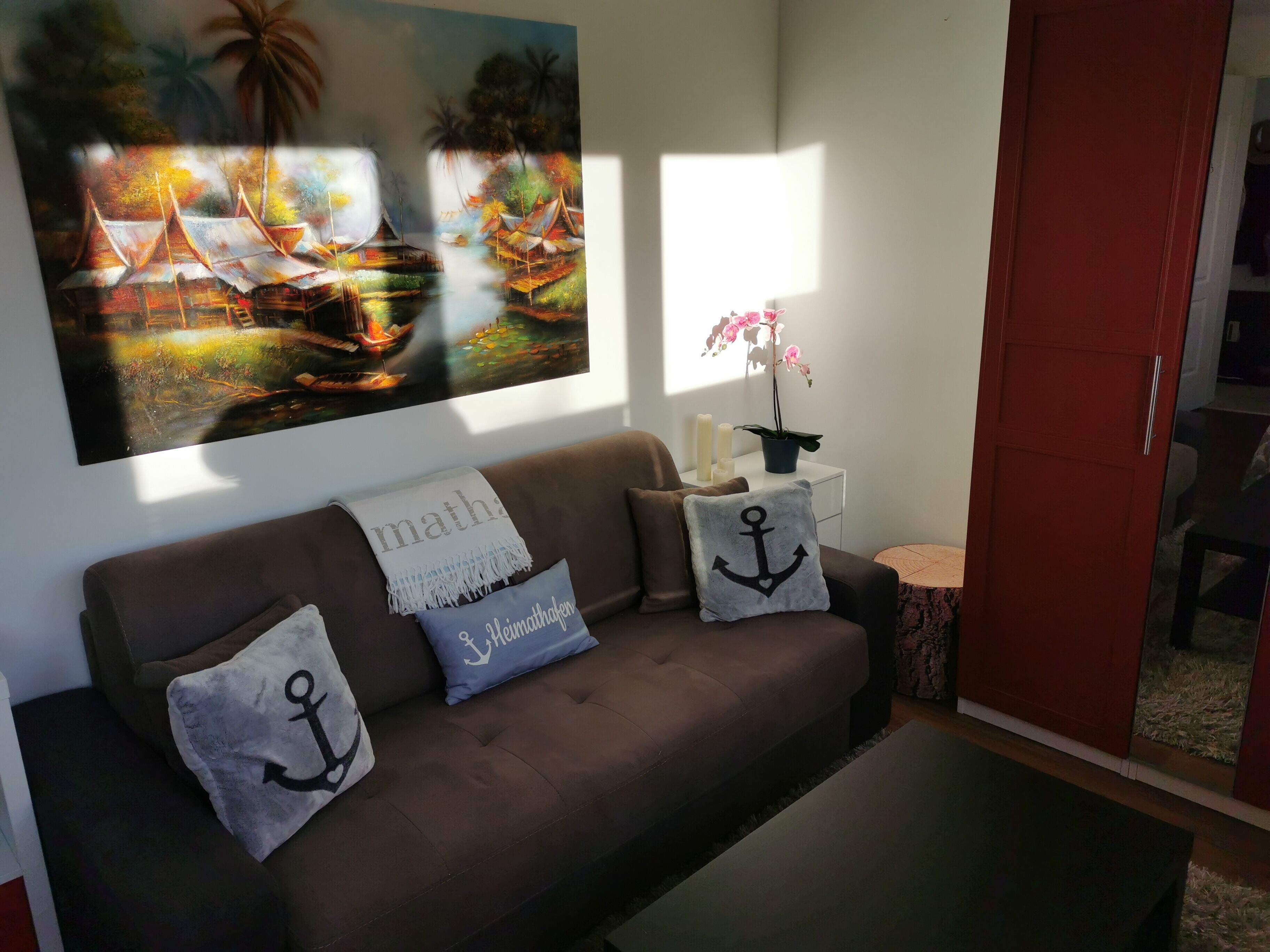 Ein Wohnzimmer mit einem braunen Sofa, drei Kissen mit Anker-Motiven, einem Gemälde mit einer tropischen Szene sowie einem kleinen Tisch mit Kerzen und einer Topfpflanze.