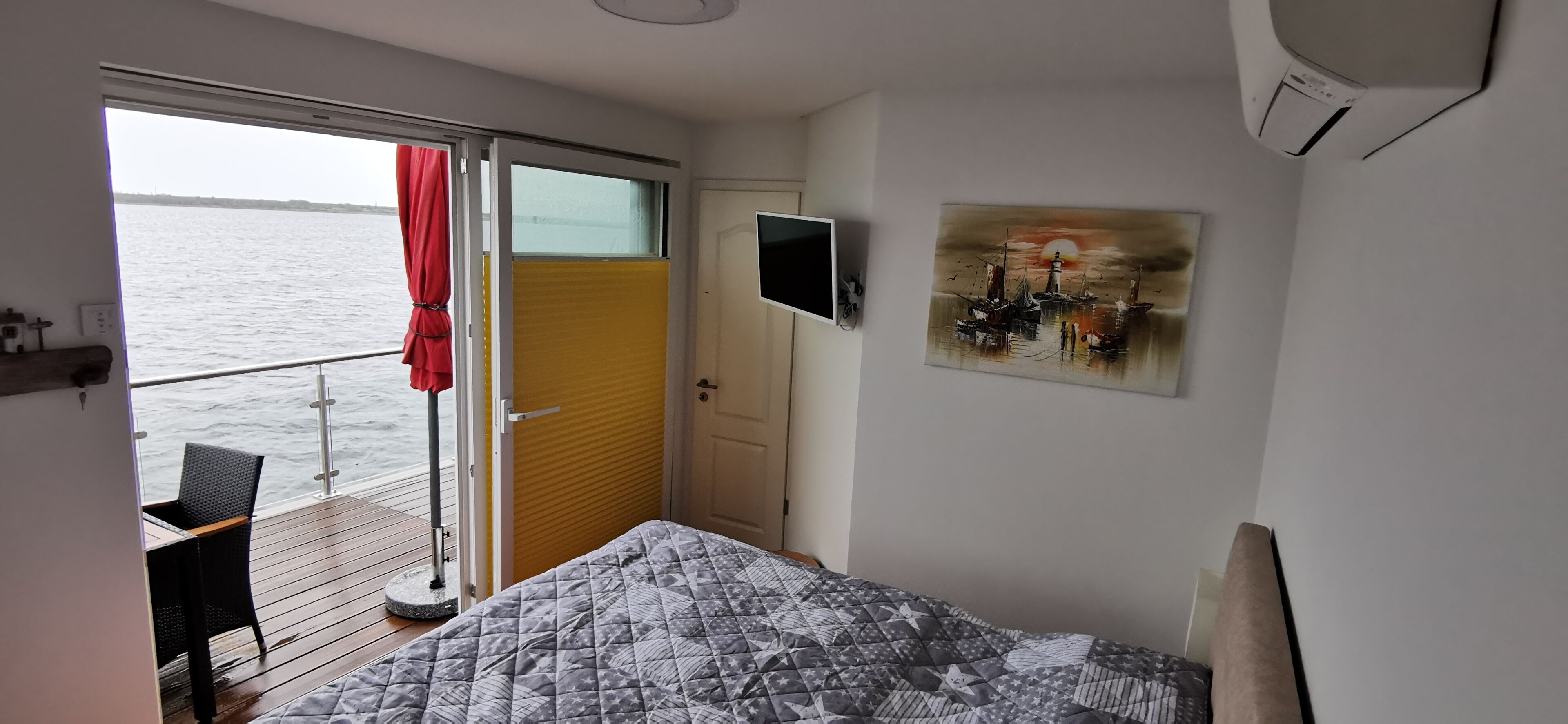 A bedroom with a bed, a TV, a painting, and a view of the water through a sliding door.