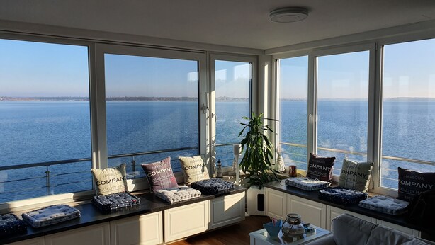 A room with large windows overlooking a body of water, featuring a seating area with cushions and a plant.