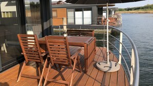 Una terraza de madera con una mesa, sillas, barandilla metálica y un gran paraguas.