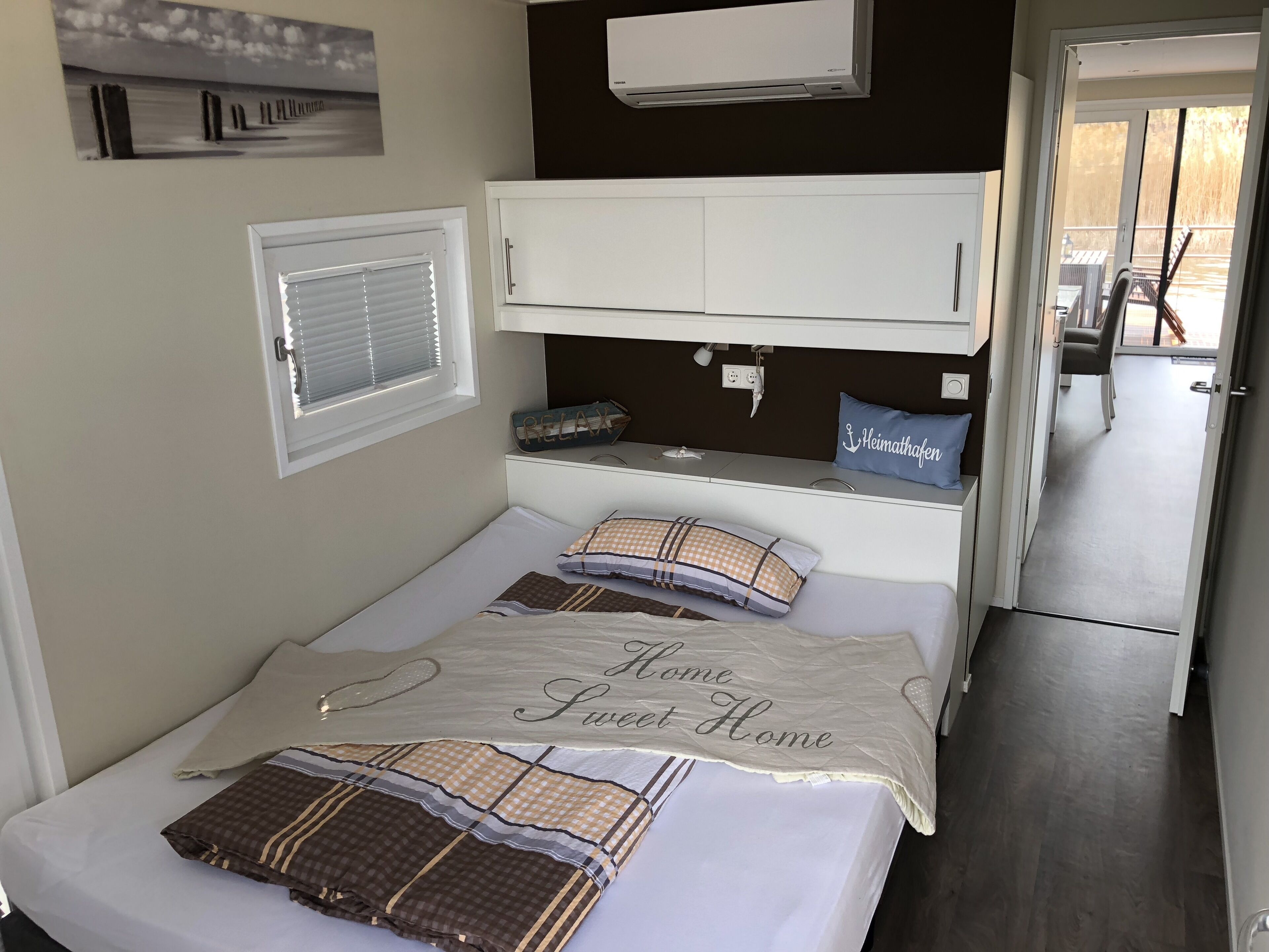 Ein Schlafzimmer mit einem Bett, einem Fenster mit Jalousien, einem weißen Kleiderschrank, einem blauen Kopfkissen und Blick auf den Balkon.