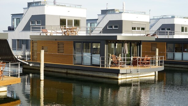 Una casa flotante moderna con terraza, ventanas grandes y una masa de agua al fondo.