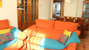 A living room with an orange sofa, blue and multicolored pillows, a wooden cabinet, and a dining area with a table and chairs.