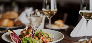 Une table à manger avec une assiette de nourriture, deux verres de vin blanc et un panier de pain.