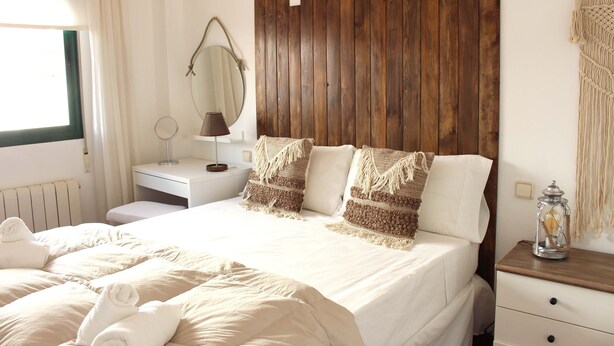 A neatly made bed with white linens and two decorative pillows, a bedside table with a lamp, a wooden headboard, and a small nightstand with a lantern.