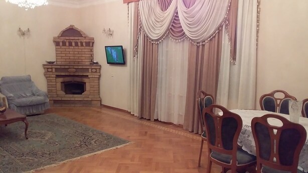 A living room with a brick fireplace, a television mounted on the wall, a dining table with chairs, and a blue armchair. - Family Apartment (Baku)