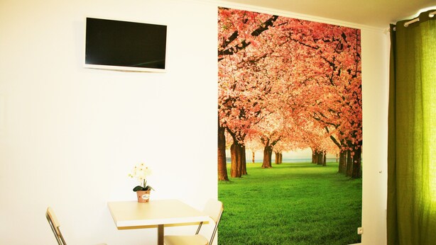 Una habitación con pared blanca, un televisor de pantalla plana montado sobre una pequeña mesa con dos sillas, y un ventanal con cortina verde.