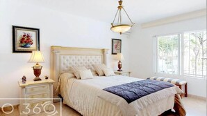 A bedroom with a large bed, two bedside tables with lamps, a window with a view of greenery, and a chandelier. - New Andalusian house (Marbella)