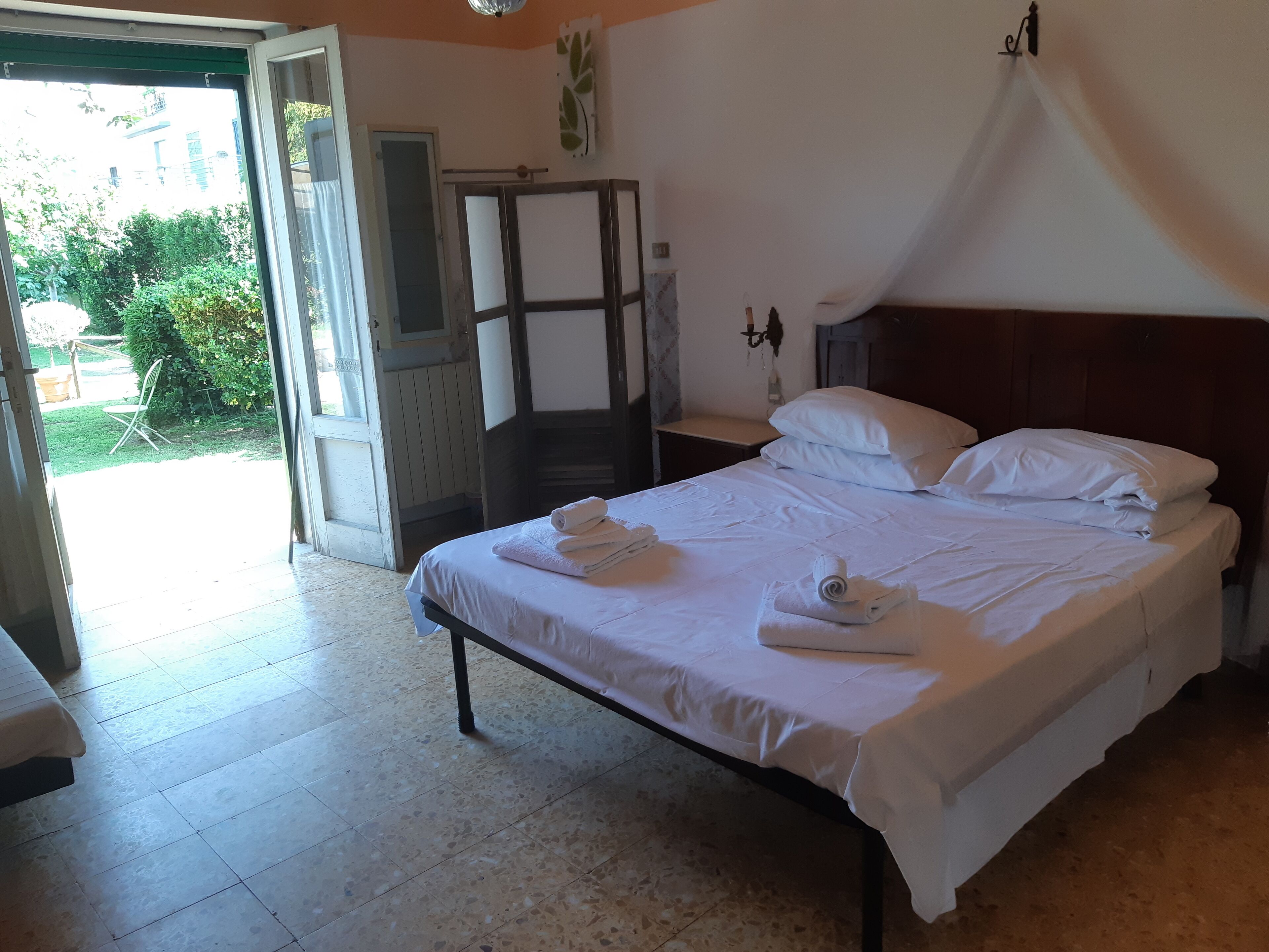 A hotel room with a bed, a nightstand, a wooden headboard, and a window with a view of greenery outside.