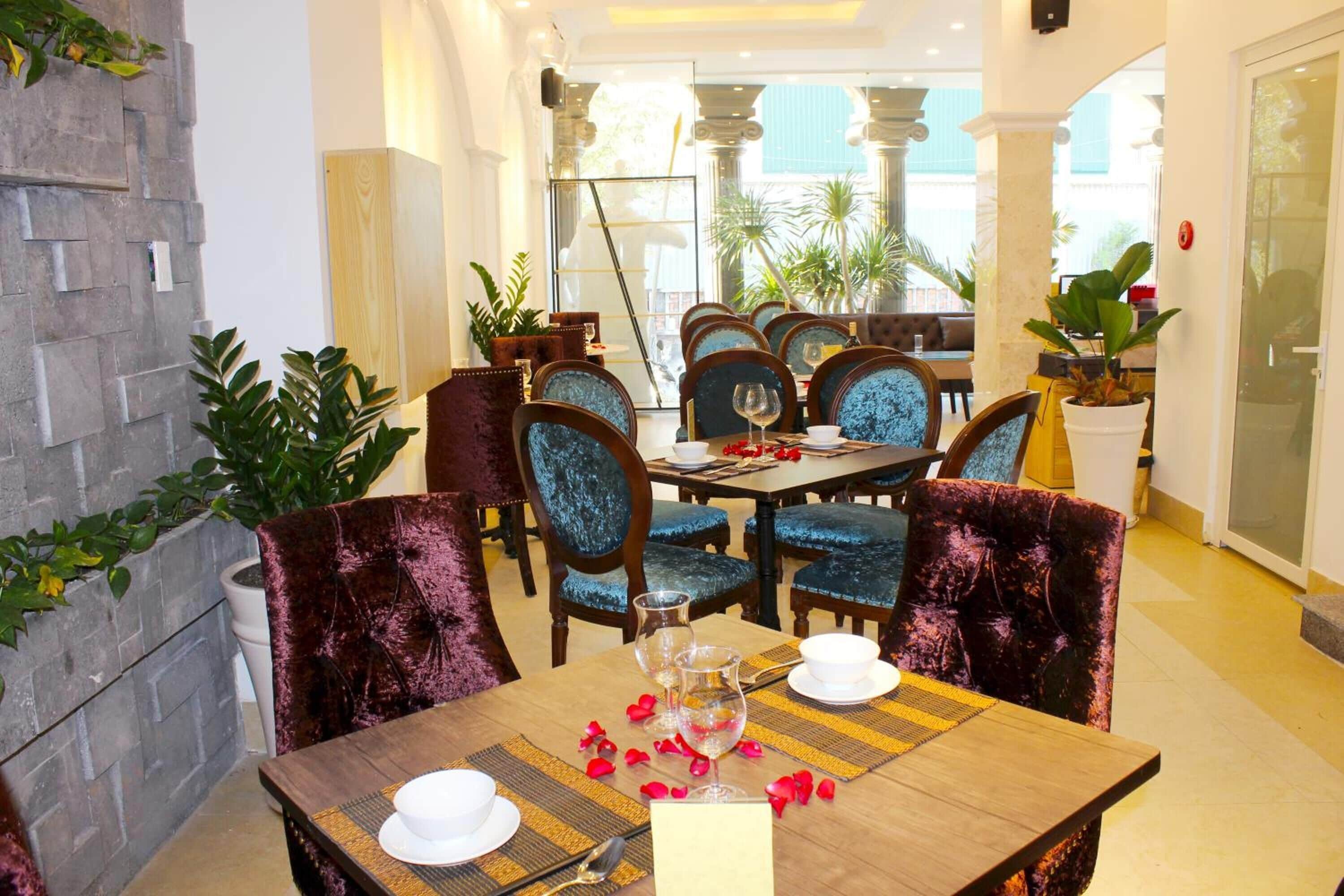 A dining area with a wooden table set for two, surrounded by velvet chairs, with a stone wall and potted plants in the background.