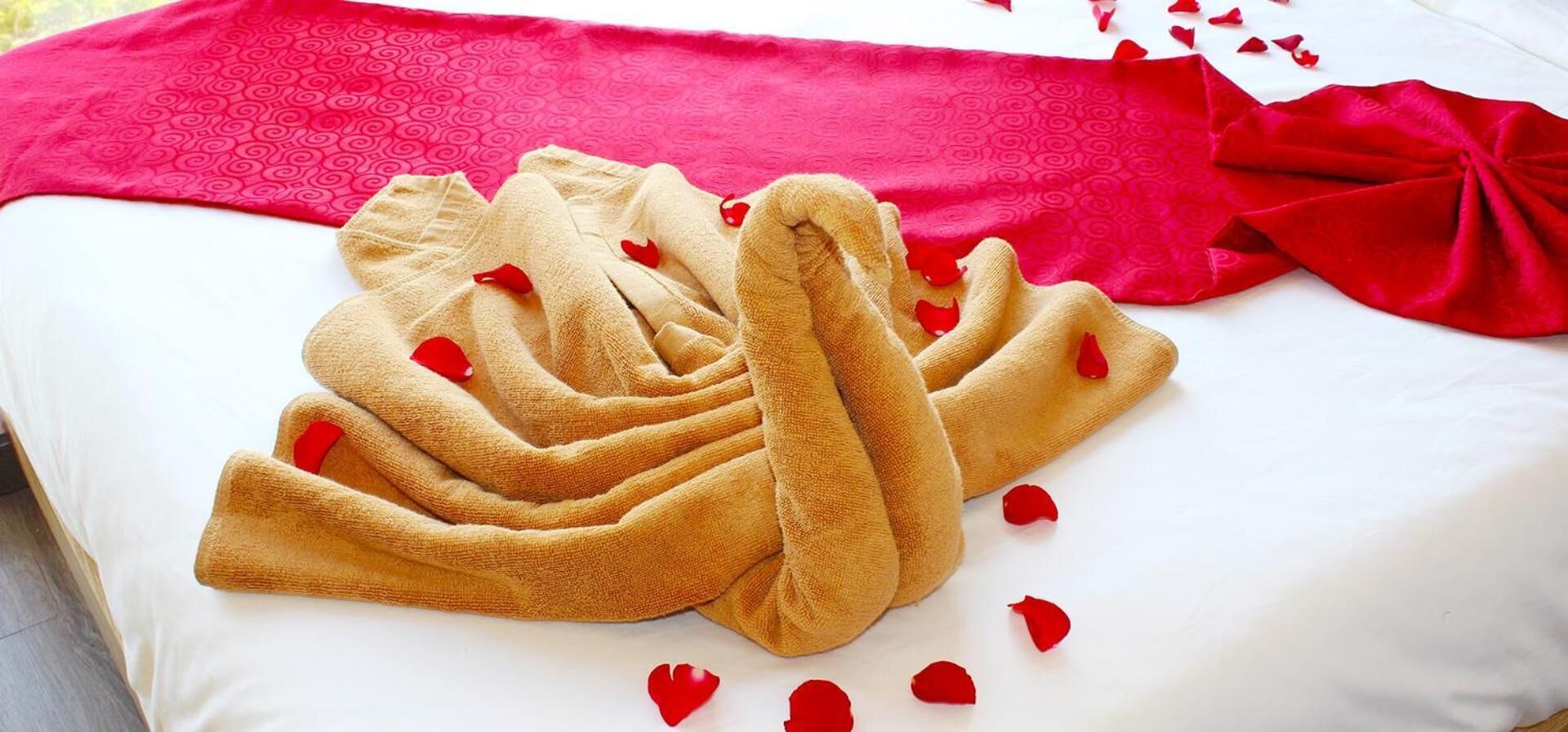 Una cama bien hecha con una manta roja y una toalla dispuesta en forma de corazón, rodeada de pétalos de rosa.
