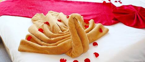 Una cama bien hecha con una manta roja y una toalla dispuesta en forma de corazón, rodeada de pétalos de rosa.