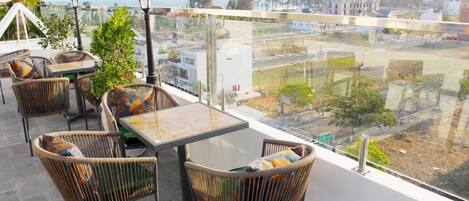 Un balcón con mobiliario de mimbre y una mesa de vidrio, con vistas a un paisaje urbano que incluye edificios y una carretera.
