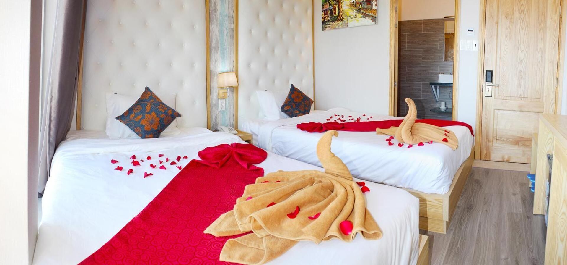 A hotel room with two beds, each with a white duvet, red and beige towels, and scattered rose petals.