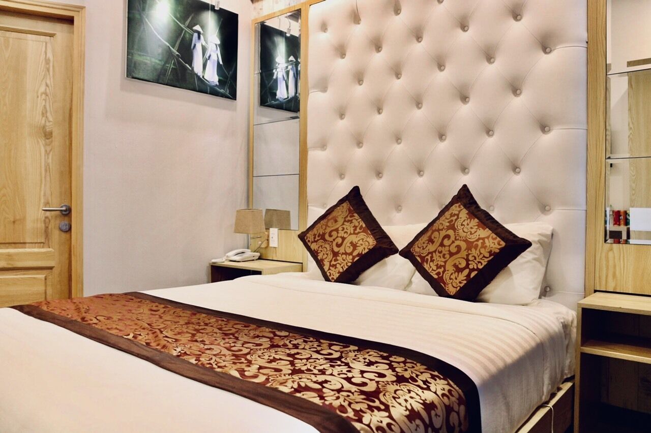 A hotel room with a tufted headboard, a large bed with patterned bedding, a bedside table with a lamp, and a mirror on the wall.