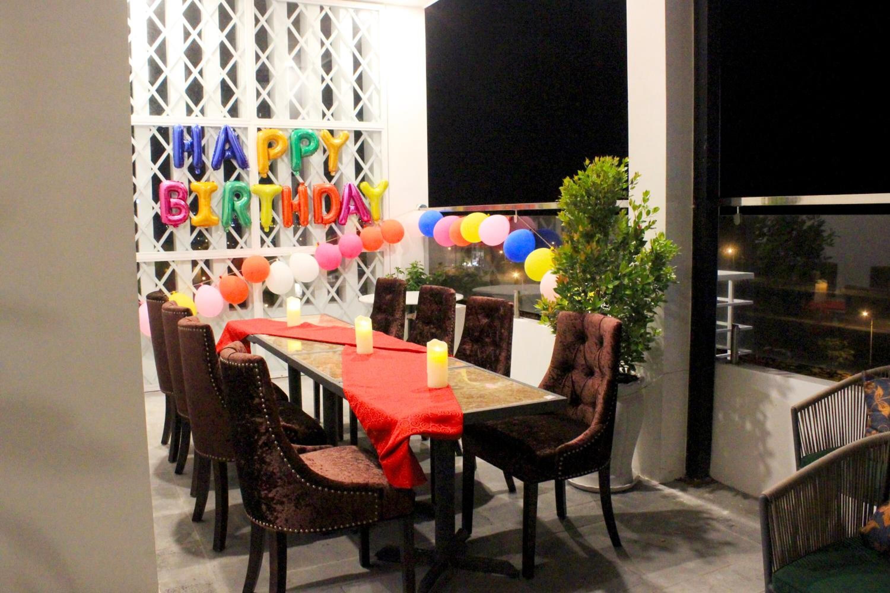A dining area with a table set for six, surrounded by velvet chairs, a red tablecloth, and a 'Happy Birthday' balloon decoration.