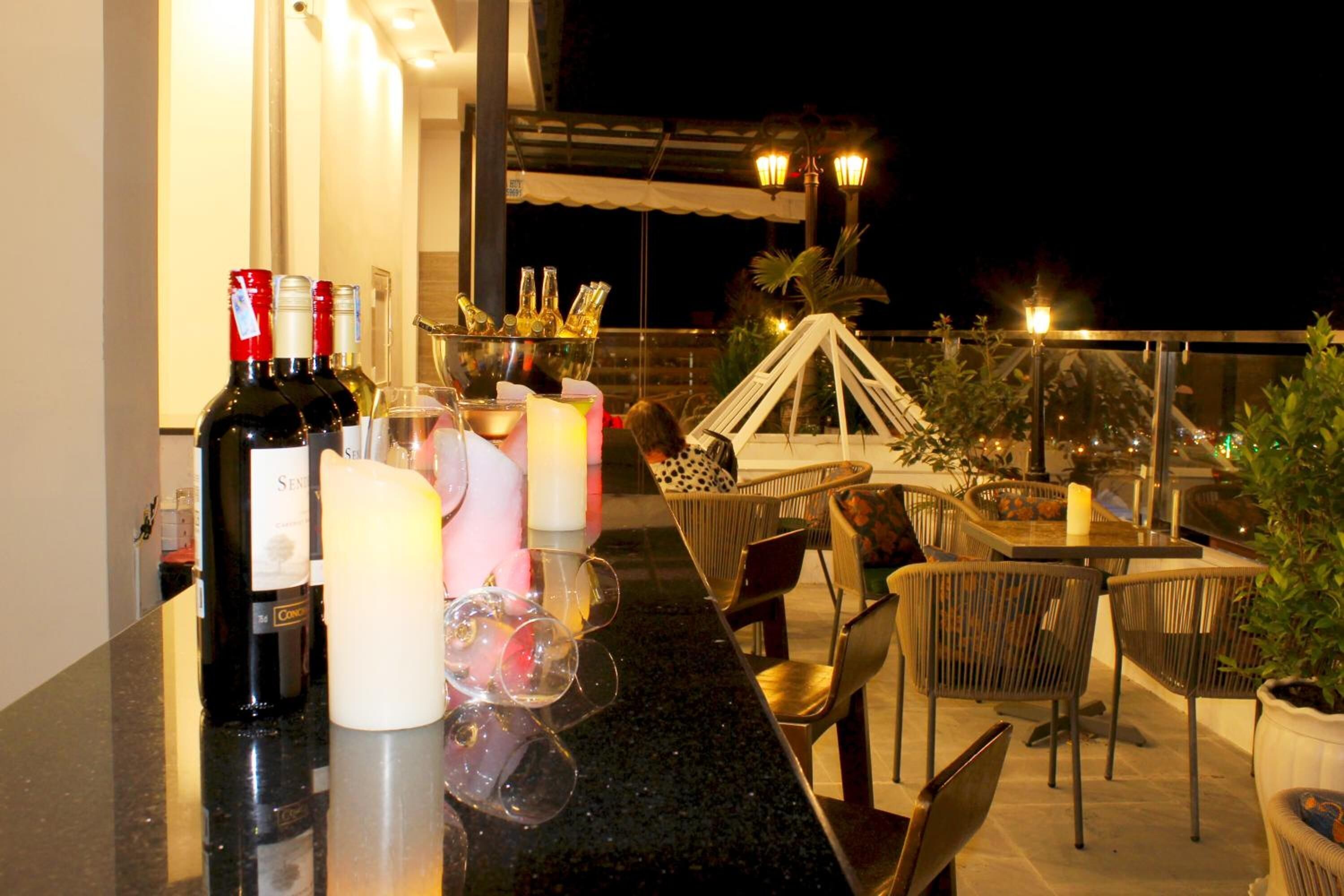 Un bar en la azotea con mostrador, botellas de vino, copas y velas. Hay sillas de mimbre y una mesa con vistas a la ciudad iluminada de noche.