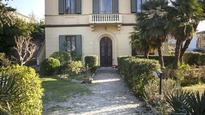 A two-story building with a symmetrical facade, a central entrance with an arched doorway, and a gravel pathway leading to the entrance. - Butterfly house. Apartment in Villa. Shared swimming pool and large park (Torre del Lago Puccini)
