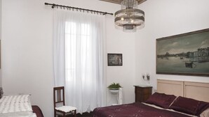 A bedroom with a large bed, a chair, a small table with a plant, a painting on the wall, and a chandelier. - Butterfly house. Apartment in Villa. Shared swimming pool and large park (Torre del Lago Puccini)