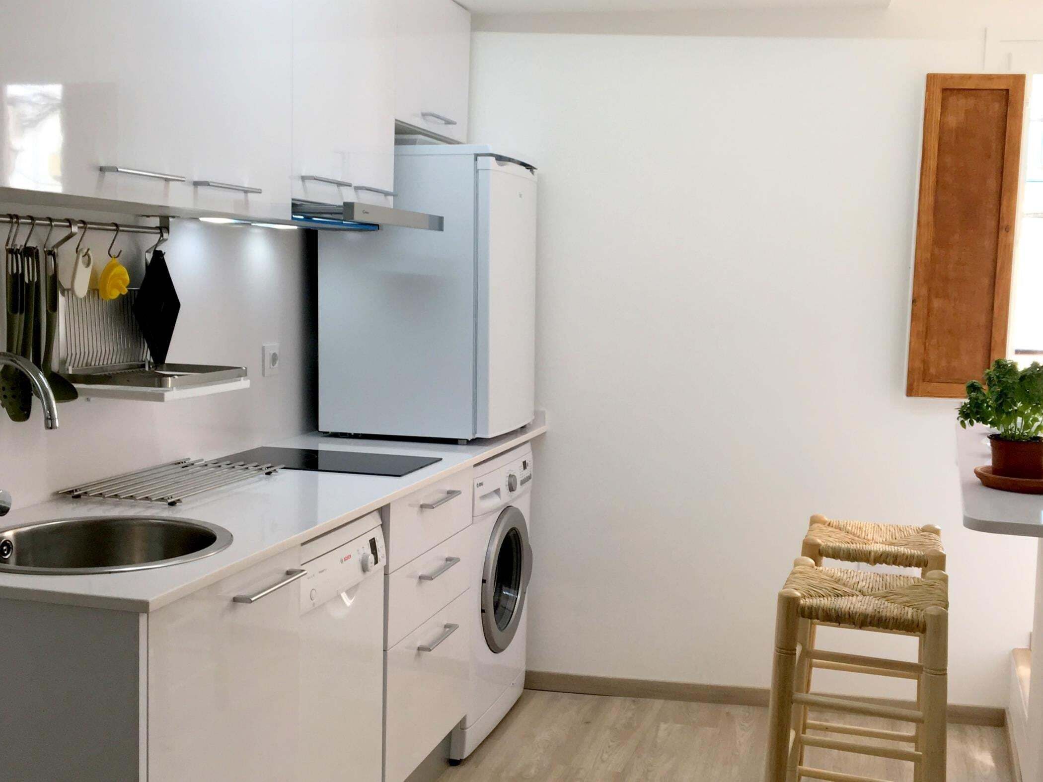 Een kleine keuken met een wasmachine, een spoelbak en een klein krukje.