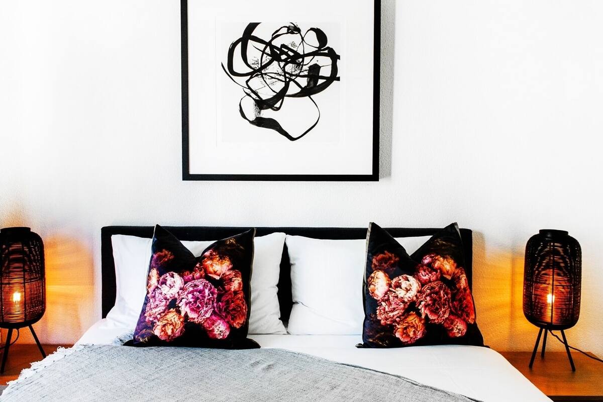 A neatly made bed with white linens and two decorative pillows with floral designs. There are two wall-mounted lamps on either side of the bed.