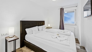 Una cama bien hecha con sábanas blancas, una mesita de noche con lámpara, una ventana con cortinas y un televisor instalado en la pared.