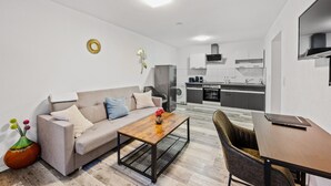 Una sala de estar moderna con un sofá, una mesa de centro y un comedor con una mesa y sillas. Se ve una cocina al fondo con un microondas, estufa y armarios.