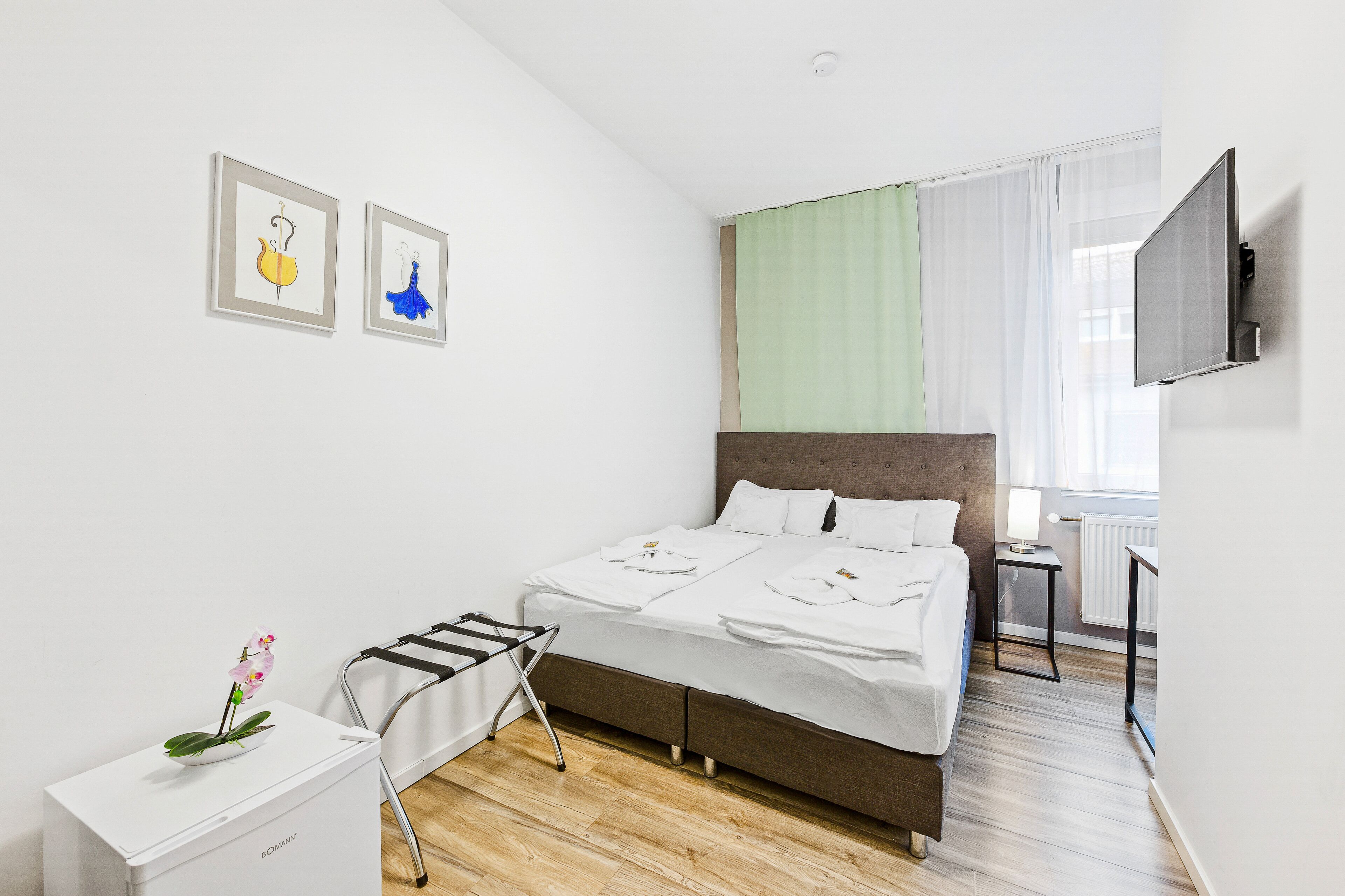 A hotel room with a bed, a TV mounted on the wall, a radiator, and a window with curtains.