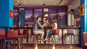 Un espace bar avec deux personnes assises au comptoir, des chaises en bois et une vue sur le paysage urbain à travers de grandes fenêtres.