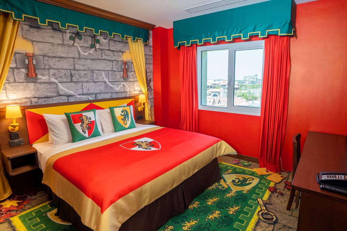 Habitación de hotel con pared de piedra, cortinas rojas y amarillas, cama con colcha roja y beige, y vista al exterior a través de una ventana.
