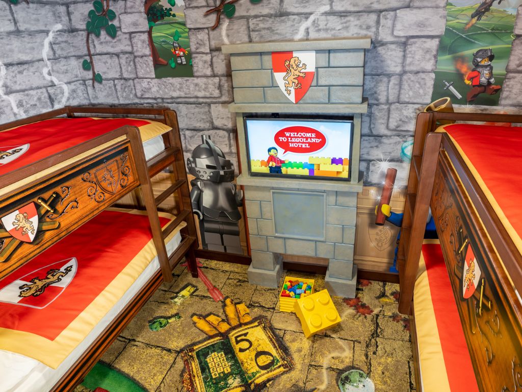A room with two wooden beds, a television screen displaying a welcome message, and a stone wall with a flag.