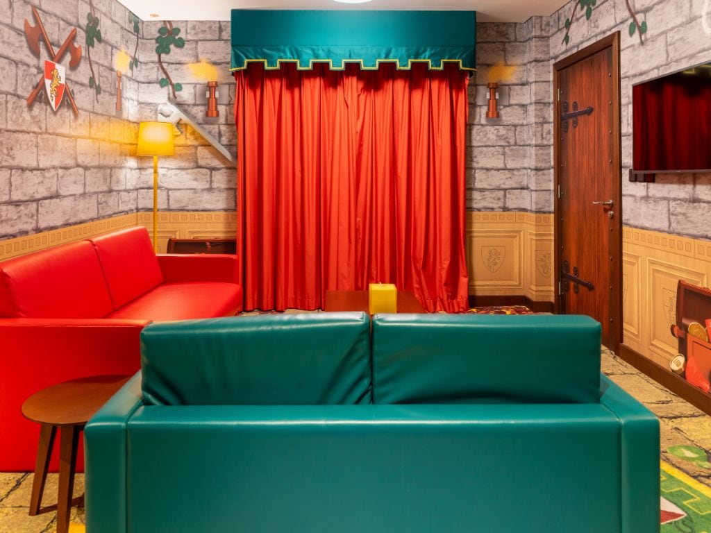 Una habitación con un sofá rojo, un sofá verde azulado, una mesita, una lámpara y una cortina.