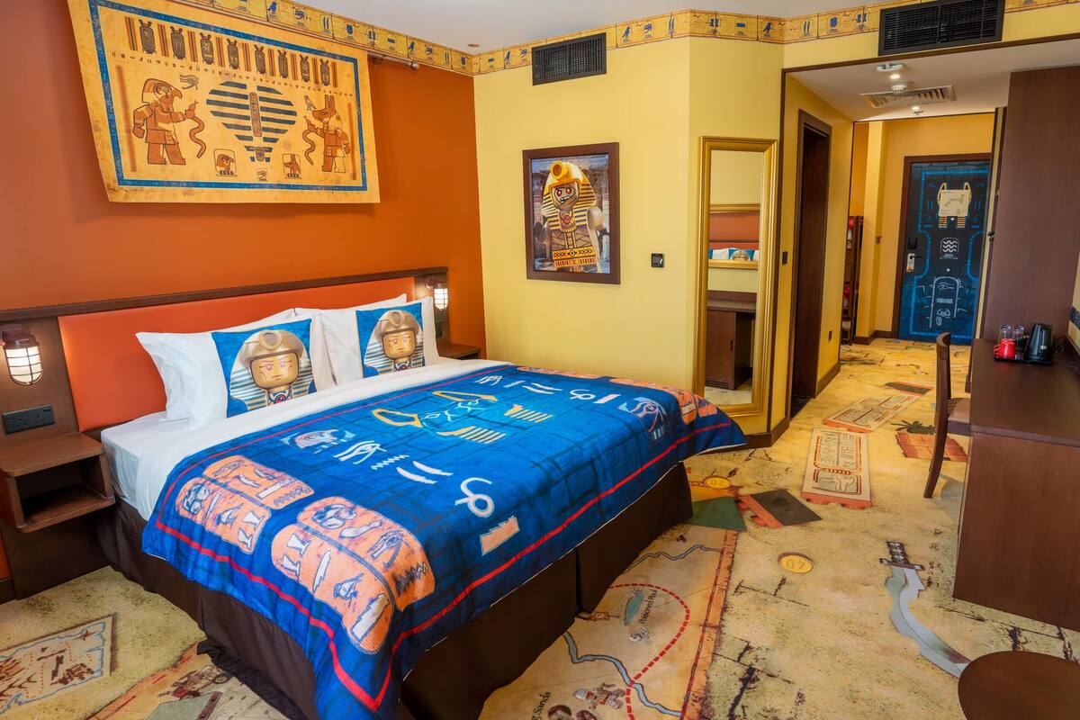 Habitación de hotel con una cama y un edredón estampado en azul y naranja, una pared con obras de arte de temática egipcia y una alfombra con diseño de mapa.