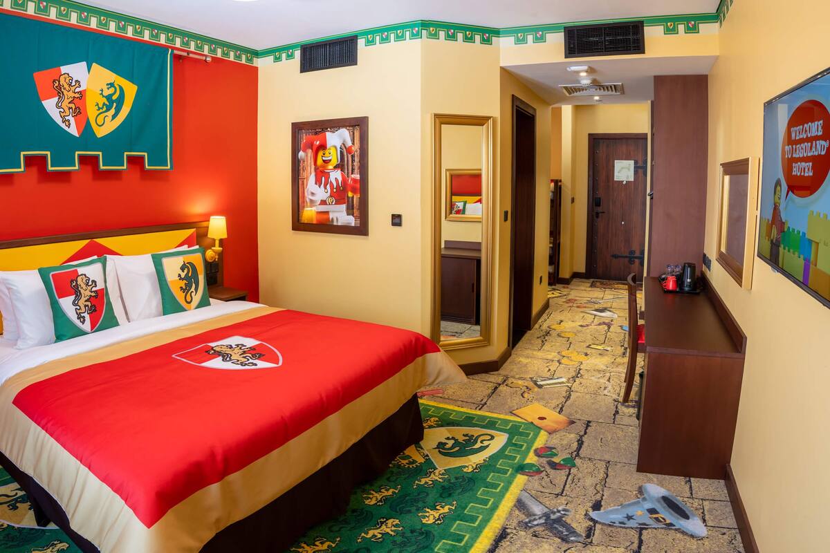 Una habitación de hotel con una cama que tiene una colcha roja y amarilla, una pared con un escudo y un pasillo con un cartel de bienvenida.