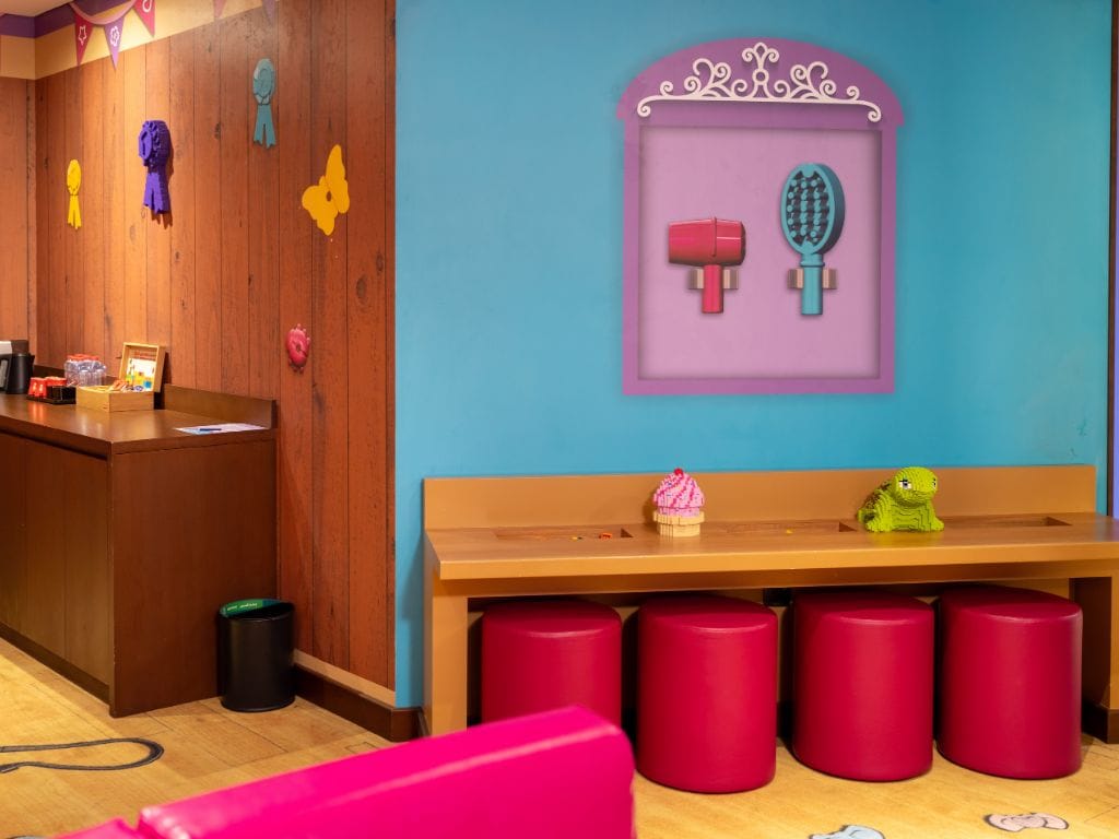 Een kinderspeelplaats met een houten bankje, rode krukjes en een muur met een lijst waarop gereedschap voor kappers staat.