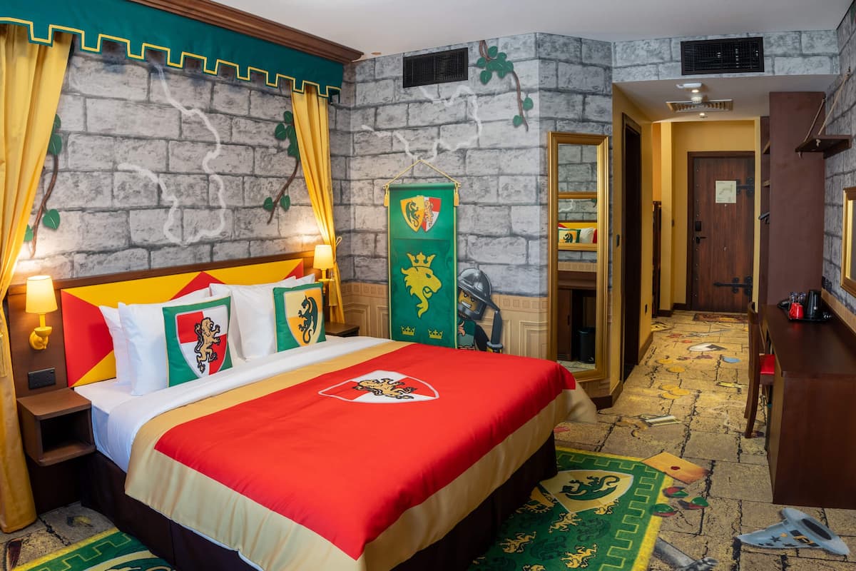 Una habitación con cama, mesita de noche, lámpara, armario, espejo, puerta y pared con un cartel que dice 'Welcome to Legoland Hotel'.