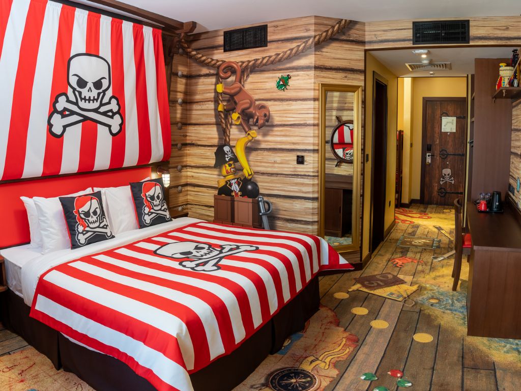 A room with a bed featuring a skull and crossbones design, a wooden wall with rope and a hanging ornament, and a door with a shield emblem.