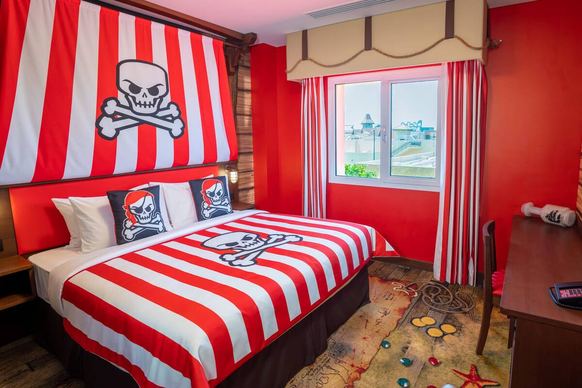 Una habitación con una cama que presenta un edredón rojo y blanco a rayas con un diseño de calavera y huesos cruzados, un cabecero de madera, un escritorio con una computadora y una ventana con vistas a edificios.
