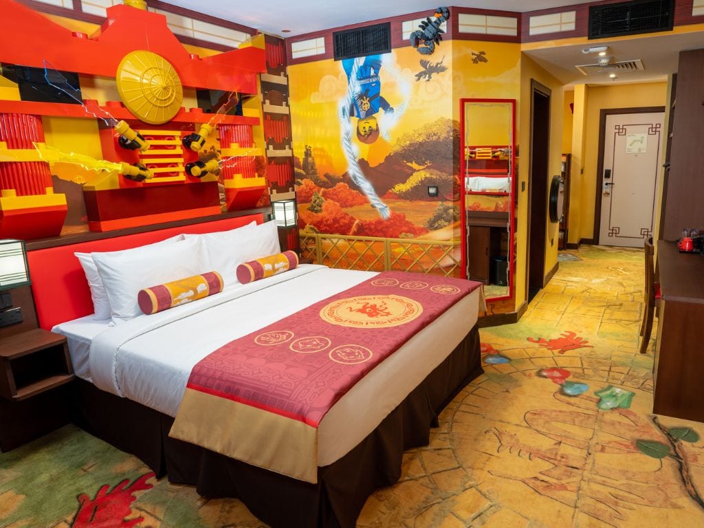 Quarto de hotel com uma cama grande, um mural na parede em tons de vermelho e amarelo, um tapete vermelho e uma porta com padrão geométrico.